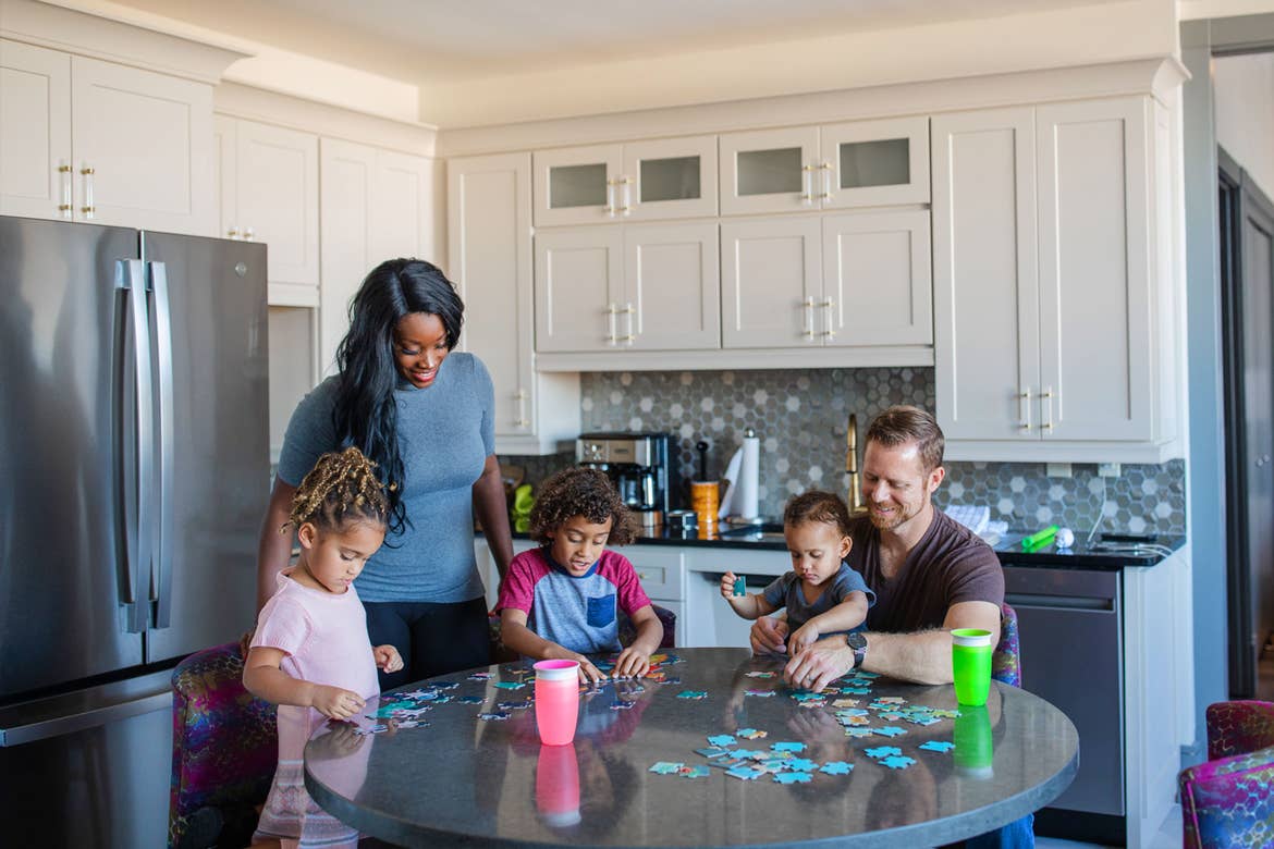 From left to right: A young girl, woman, young boy, and a dad holding a toddler sit at a kitchen table working on puzzles.