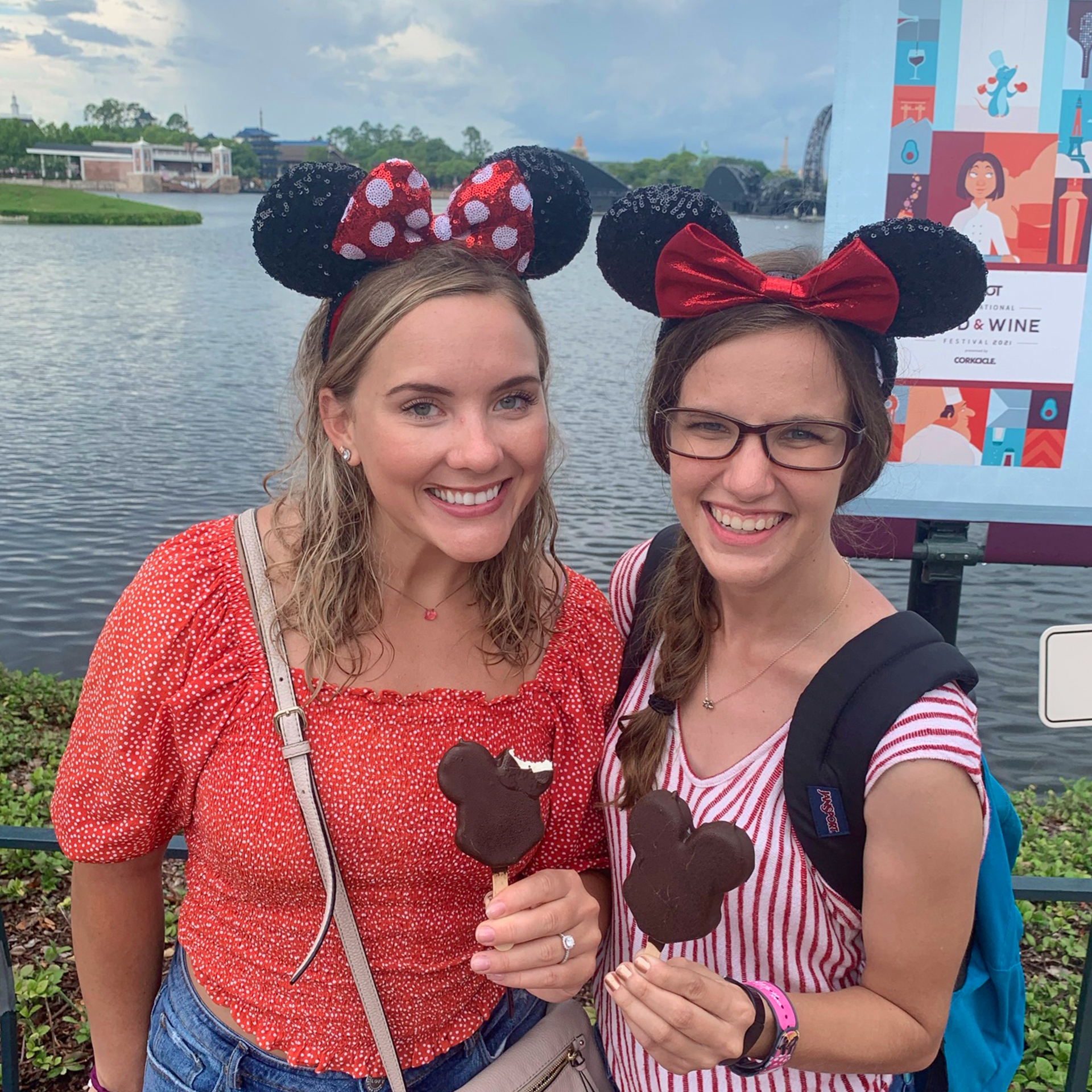Two caucasian women wearing Minnie ears stand near the World Showcase Lagoon while holding Mickey Ice Cream bars.
