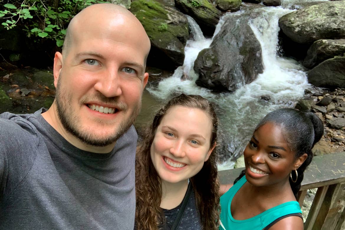 A caucasian man (left), a caucasian woman (middle) and an African American woman stand in front of the Anna Ruby Falls waterfall surrounded by lush green woods.