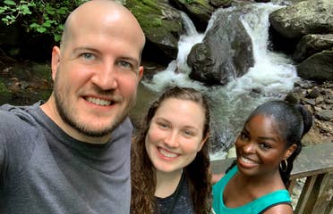 A caucasian man (left), a caucasian woman (middle) and an African American woman stand in front of the Anna Ruby Falls waterfall surrounded by lush green woods.