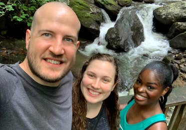 A caucasian man (left), a caucasian woman (middle) and an African American woman stand in front of the Anna Ruby Falls waterfall surrounded by lush green woods.
