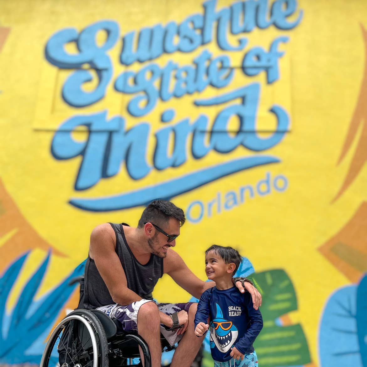 A man in a wheelchair is seated next to a toddler boy in front of a wall mural.