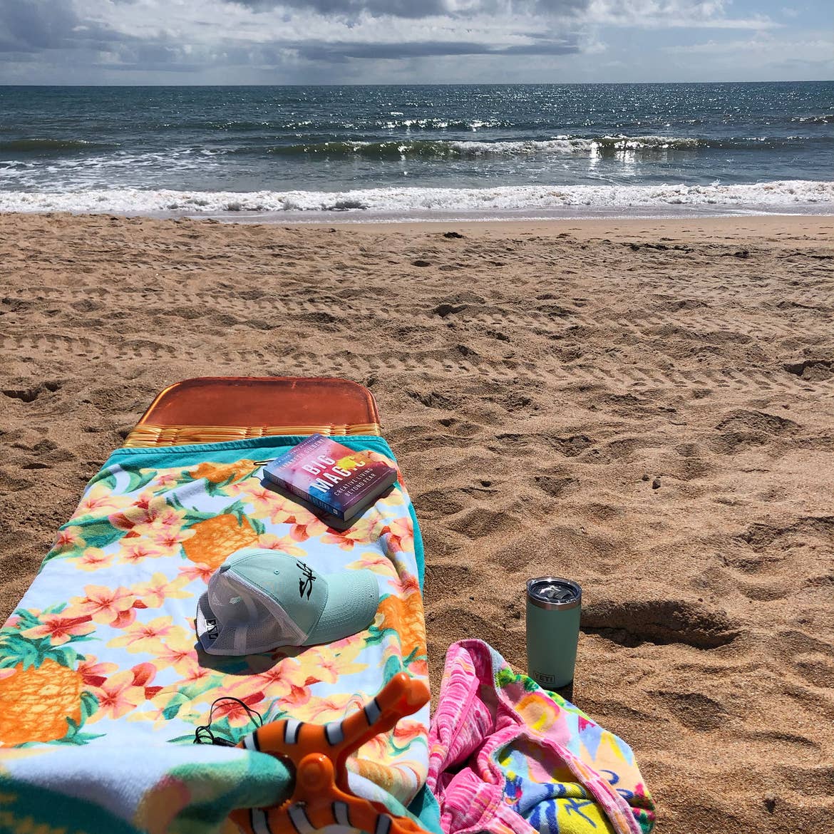 A beach chair with a baseball cap, towel, and a good book faces the ocean waves as they roll in.