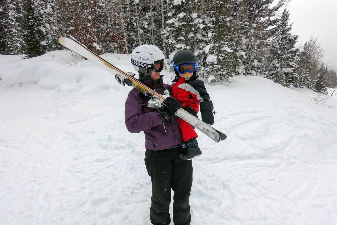 A woman holds a small child and a set of skis while they both wear winter apparel, ski helmets and goggles on a snowy slope.