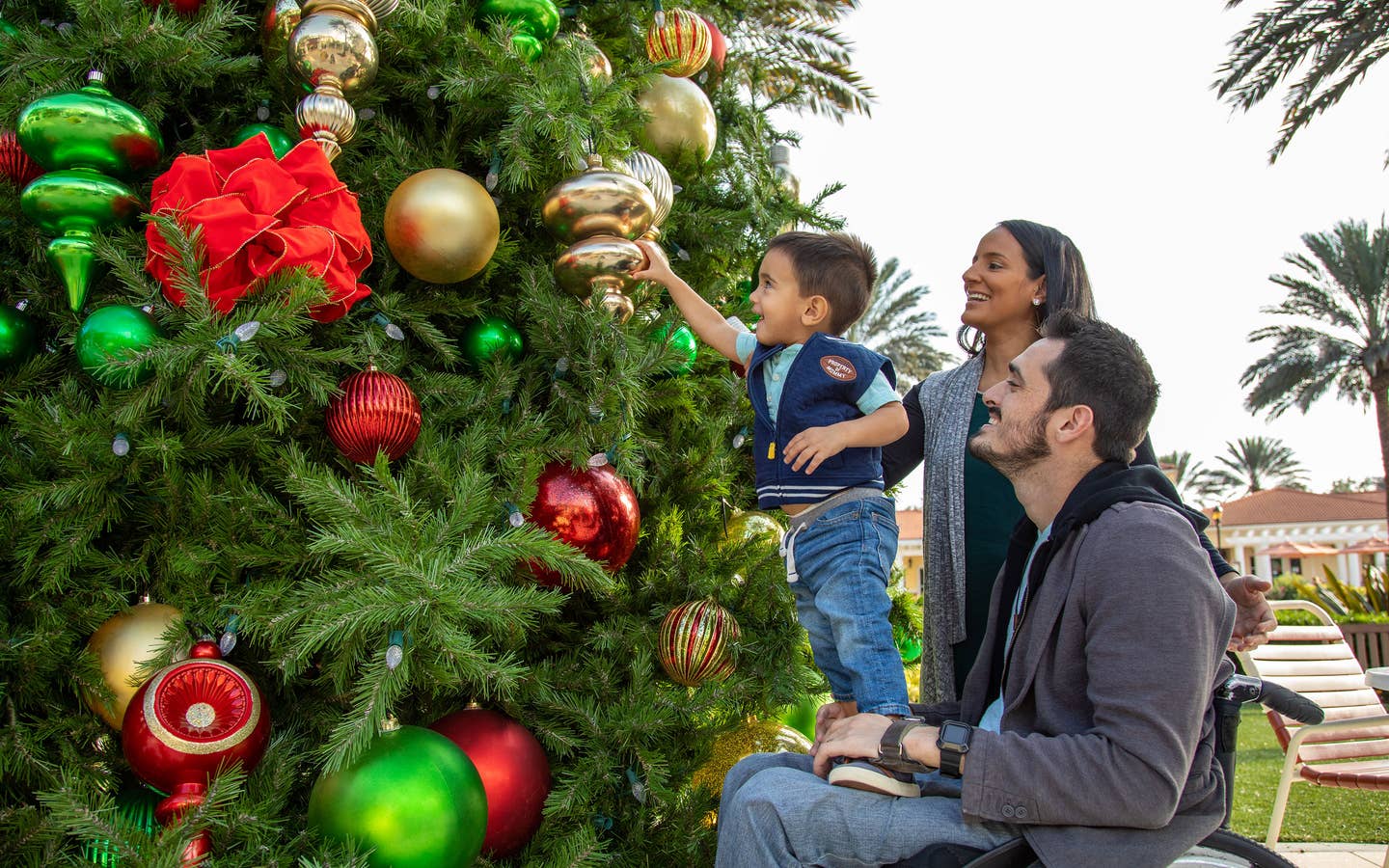 Author, Danny Pitaluga (right) sits in his black wheelchair wearing a charcoal hoodie as his wife, Val (middle), and his son, Joey (left), stands on Danny's lap to decorate a Christmas tree outdoors.