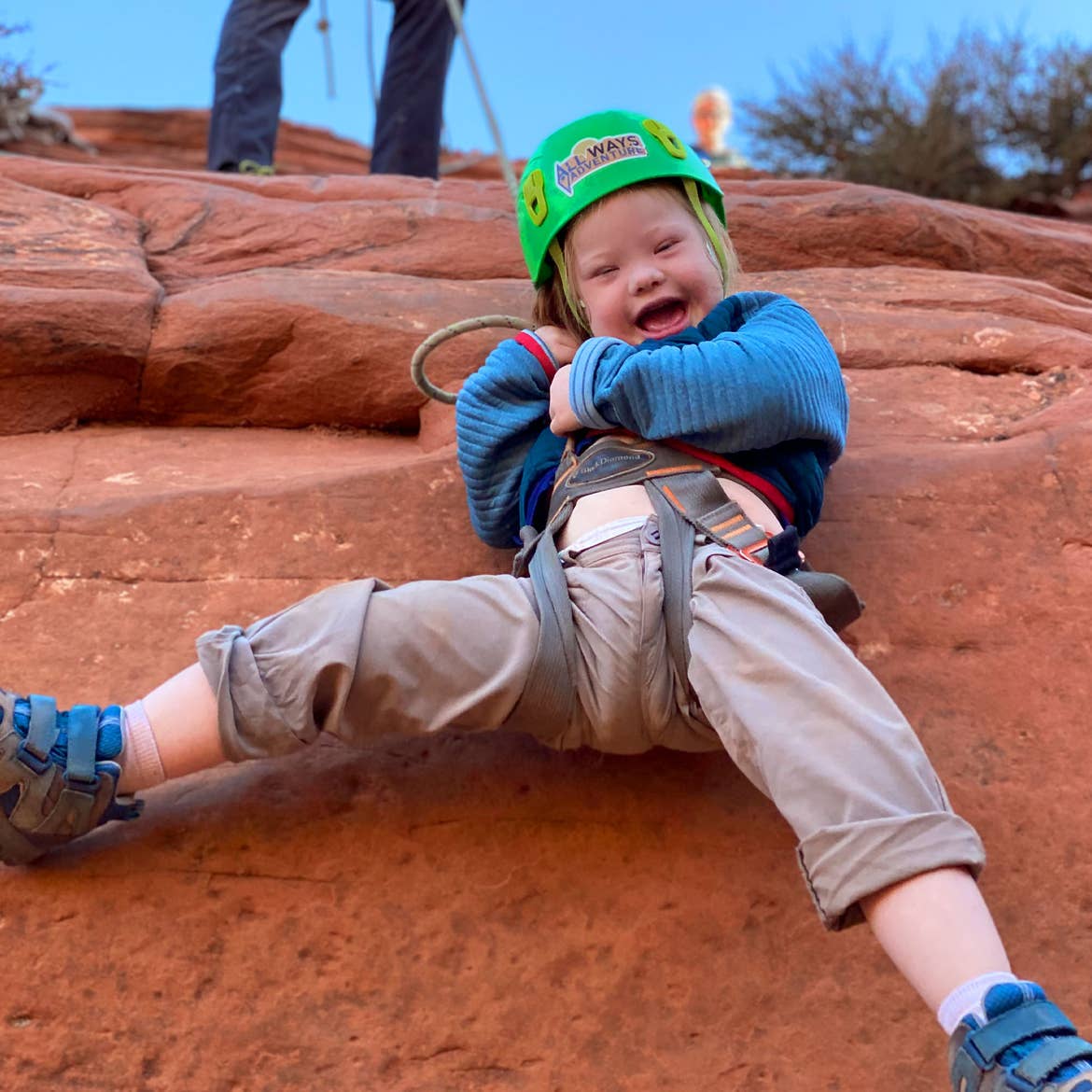 Featured Contributor, Melody Forsyth's daughter, Ruby, wears a blue jacket and green safety helmet and harness while rappelling down an orange rock formation.