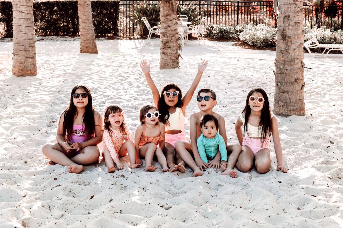Seven young children sit on the white sands wearing swimming attire and sunglasses.