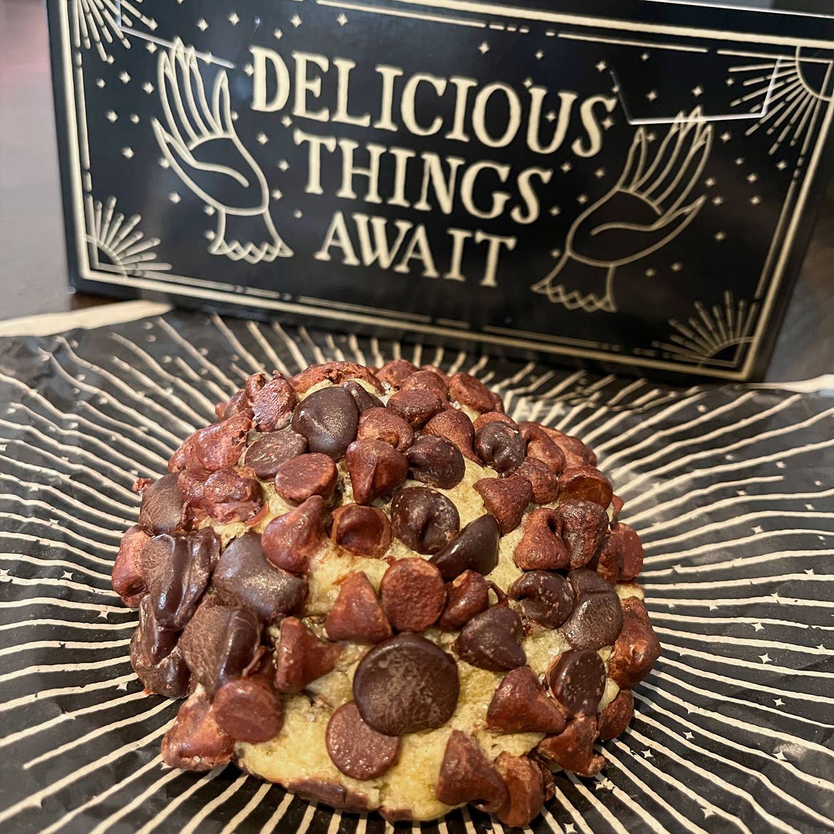 A black take-out box that reads, 'DELICIOUS THINGS AWAIT' in gold print is placed near a chocolate chip cookie on a matching wax paper.
