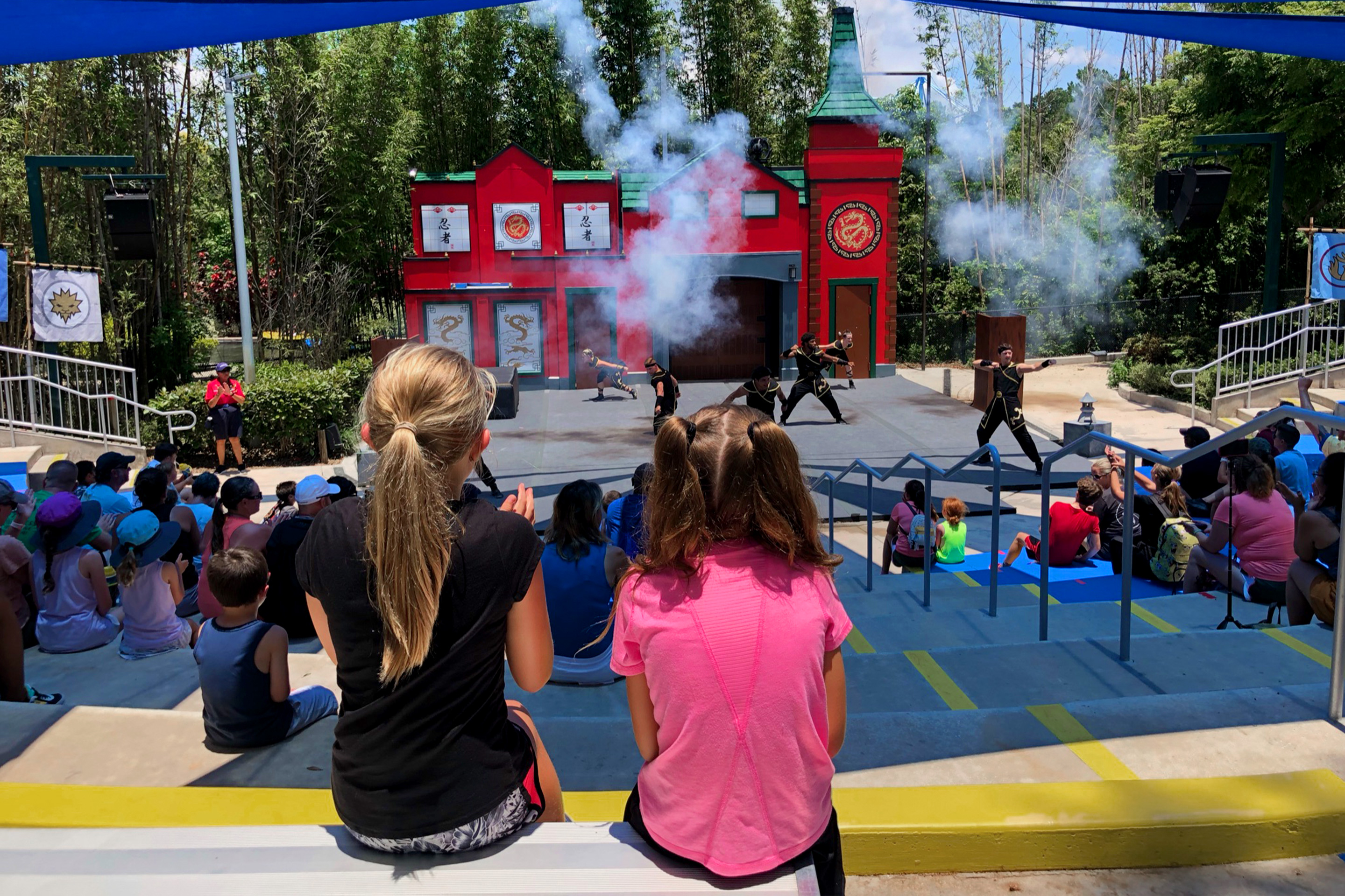 Two young girls sit in an outdoor theater watching a stage stunt show.