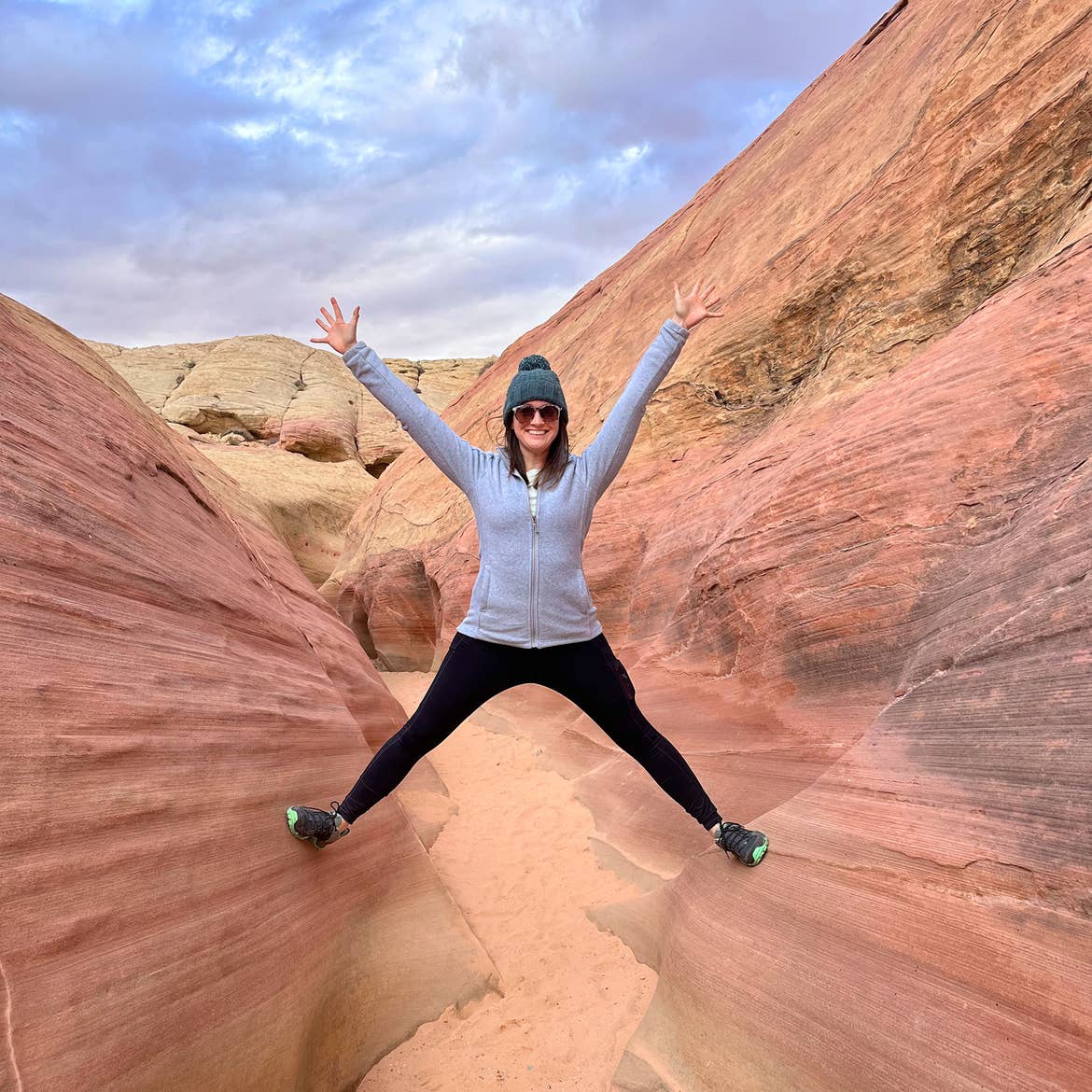 A woman wearing a grey pullover, black tights, sneakers and blue knitted cap stands between two red rock formations.