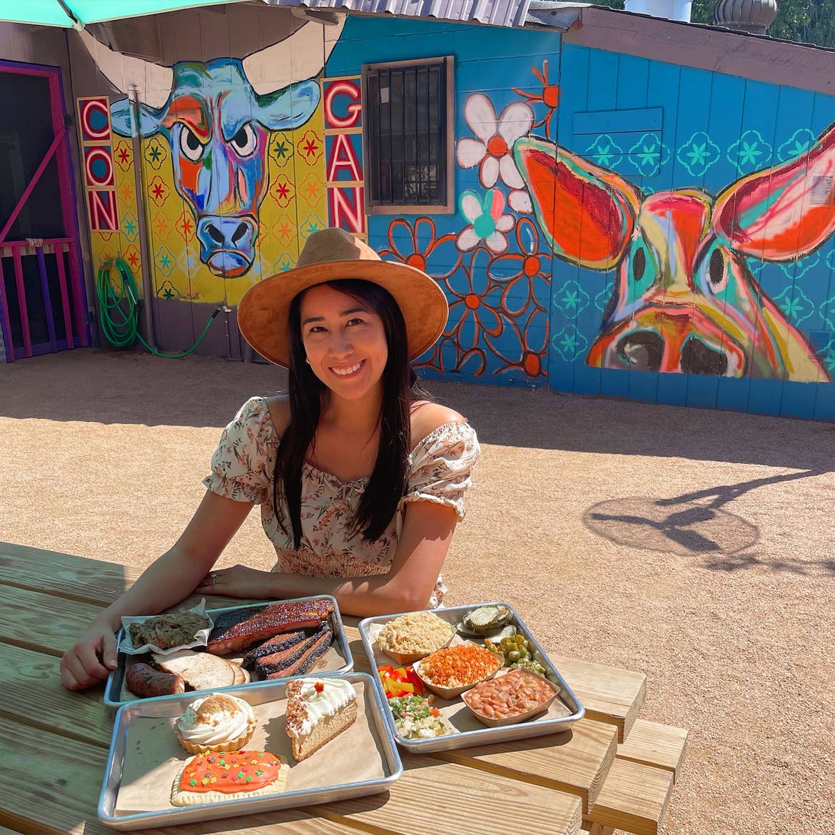 A woman in a floral sundress and sun hat sits at a wooden table with a serving tray containing barbecue dishes and sides with a painted mural of livestock behind her.