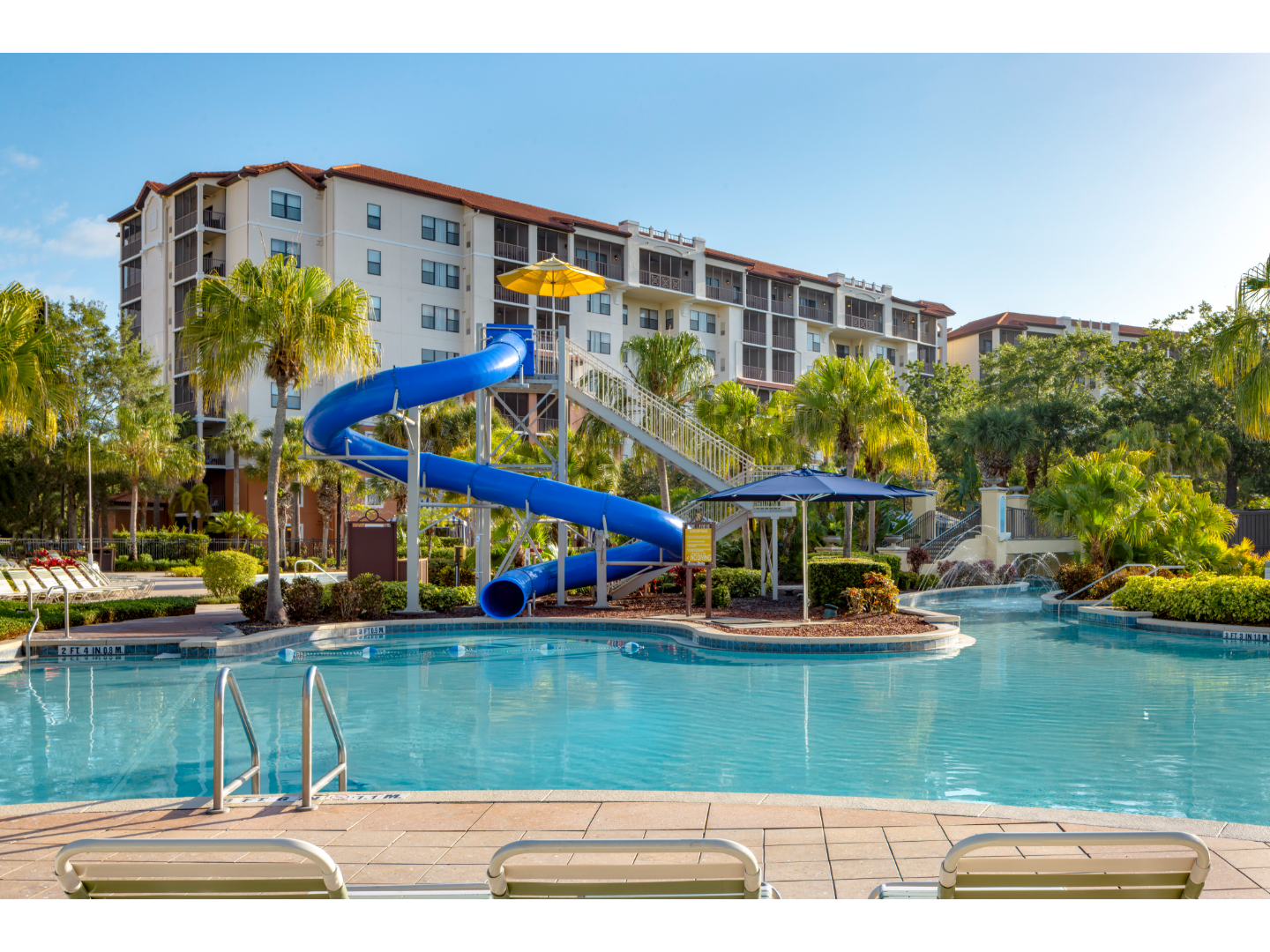 Water park pool with tall blue waterslide and resort building in background.