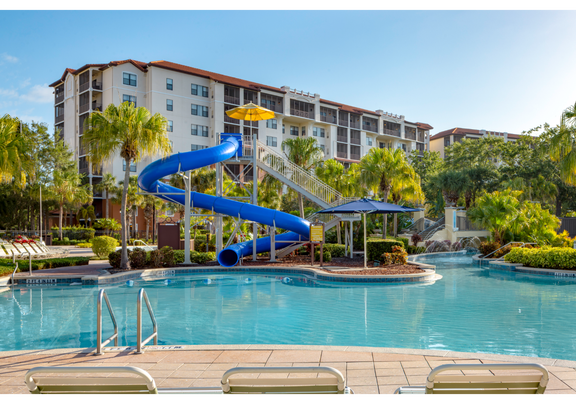 Water park pool with tall blue waterslide and resort building in background.