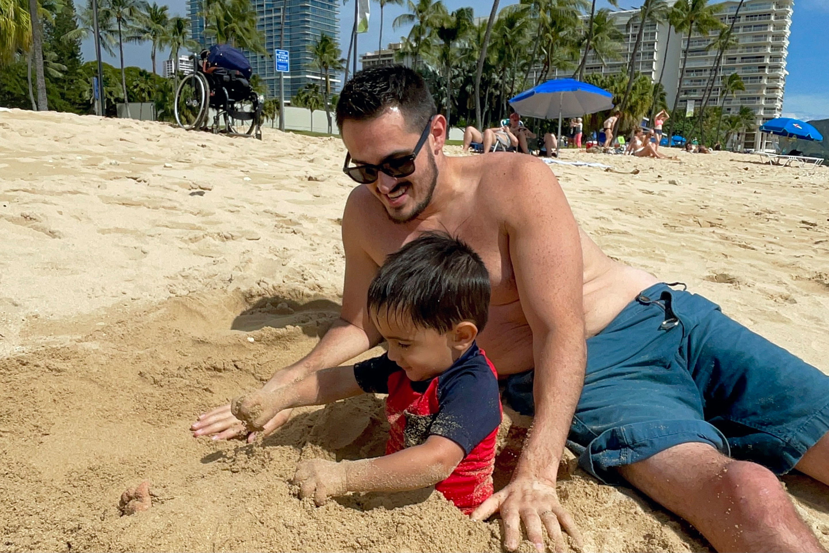 Featured Contributor, Danny Pitaluga (back) and his son, Joey (front) play with sand on the beach in swimwear.