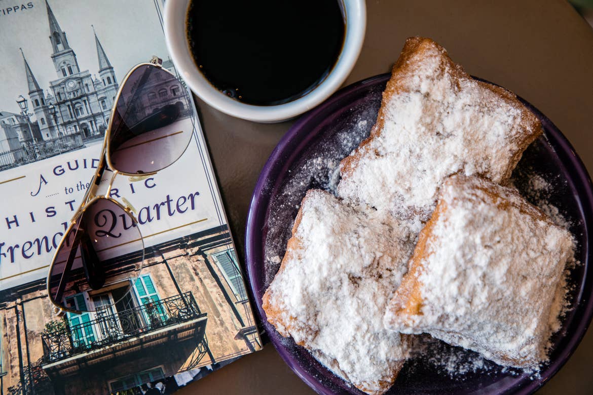 Beignets placed on a table next to a white mug full of coffee, sunglasses, and brochures that read 'A guide to the Historic French Quarter.'