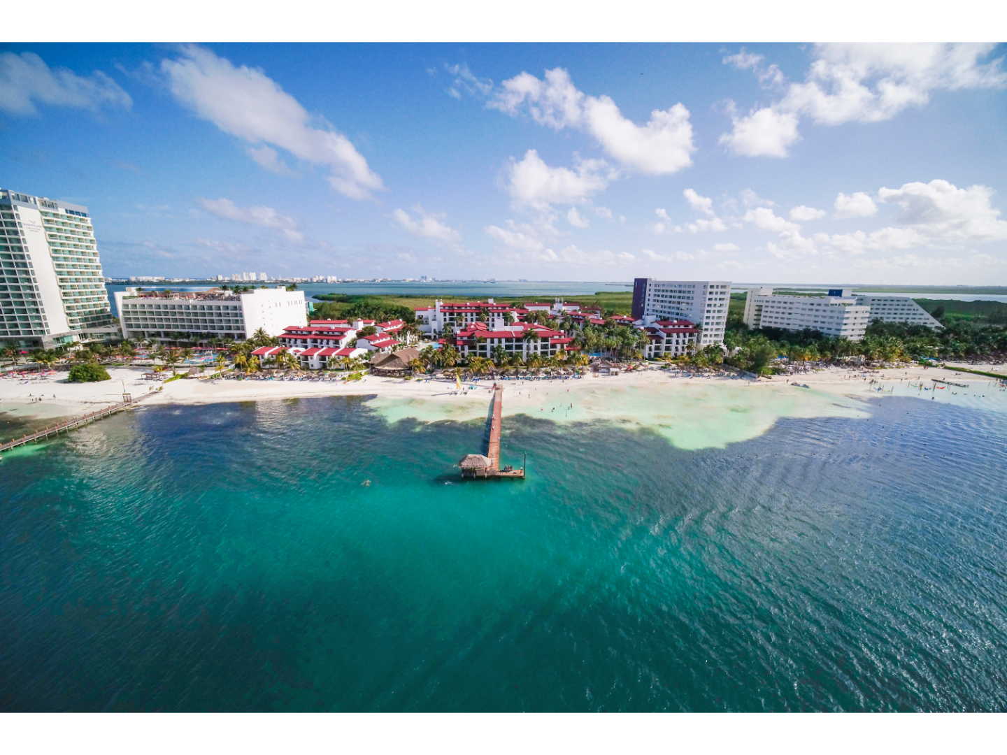Drone view of beachfront resorts with turquoise ocean water, sandy beach, and pier extending into the sea.