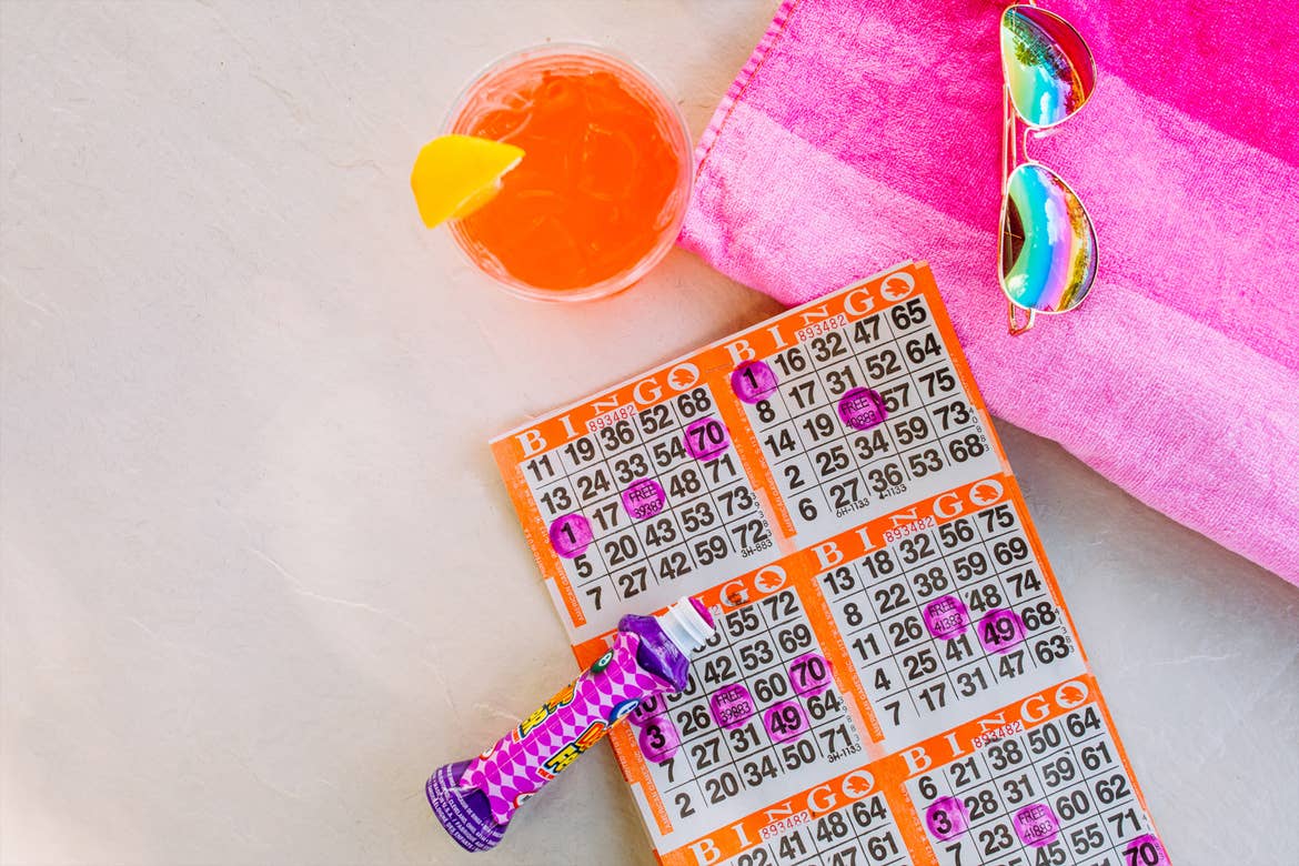 An orange and white bingo card placed on a whit table with a purple marks, sunglasses and a beverage.