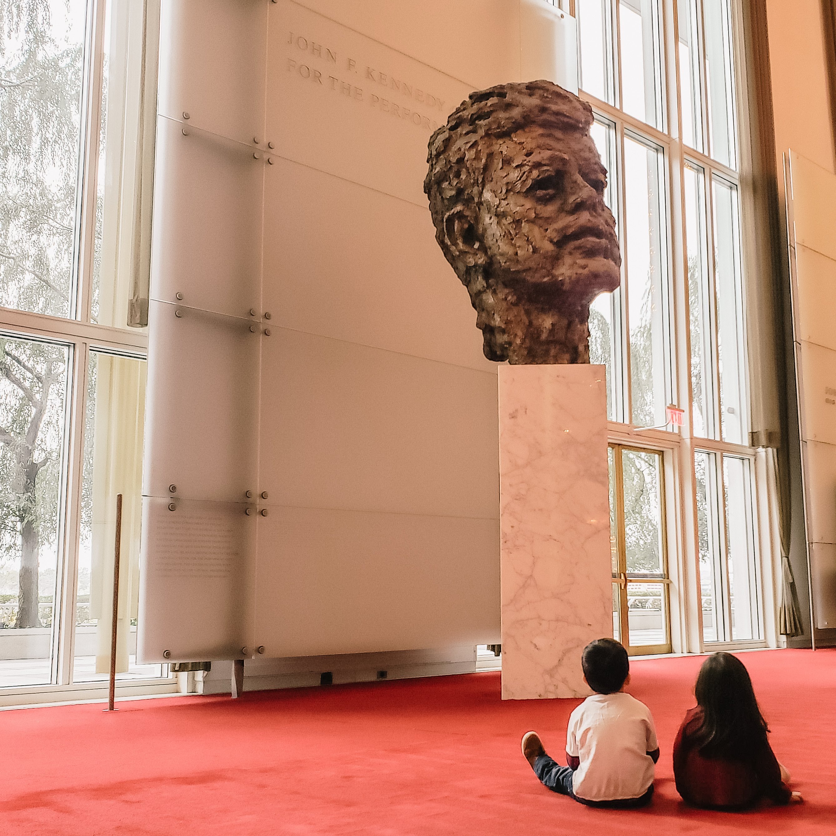 Angelica's kids sitting on the floor looking up at a statue of John F. Kennedy at the Kennedy Center.