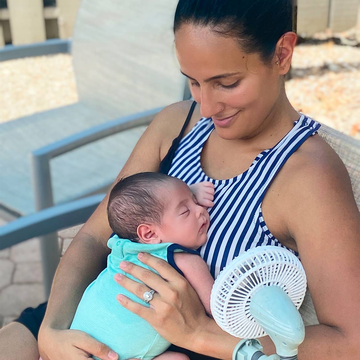 A woman sits in a chair while holding an infant near a travel fan.
