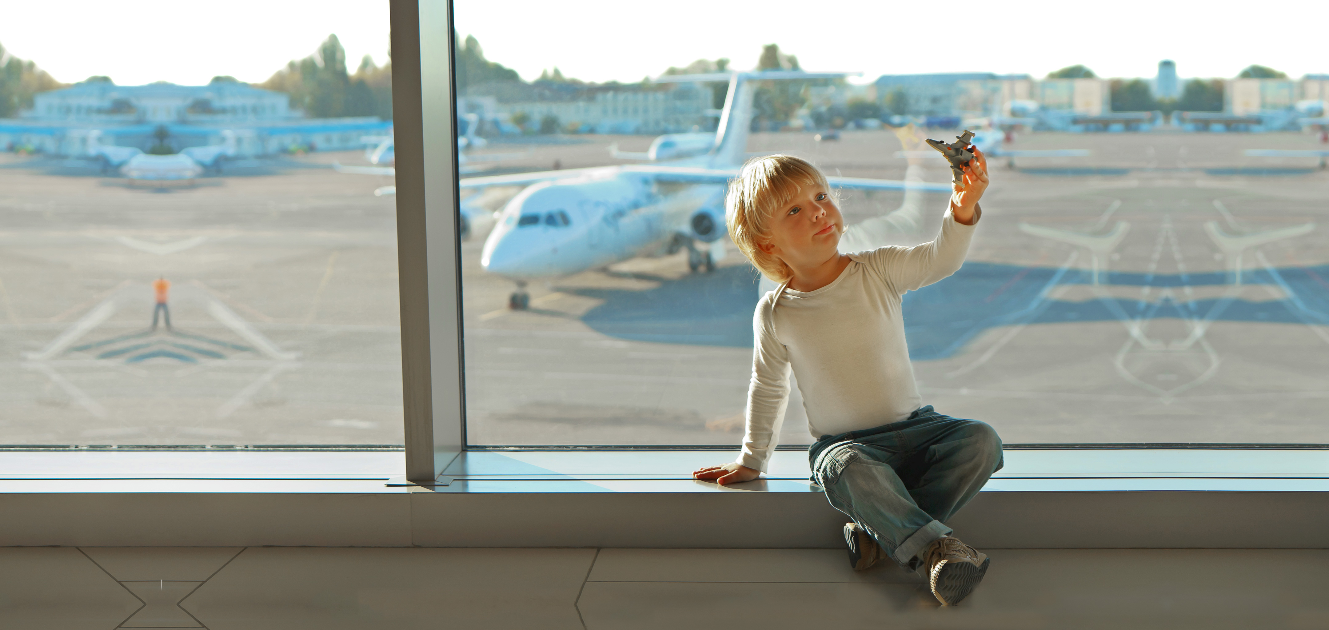 A young boy wearing a white long sleeve shirt plays with a toy airplane in front of an airport window near the runway with a white airplane.