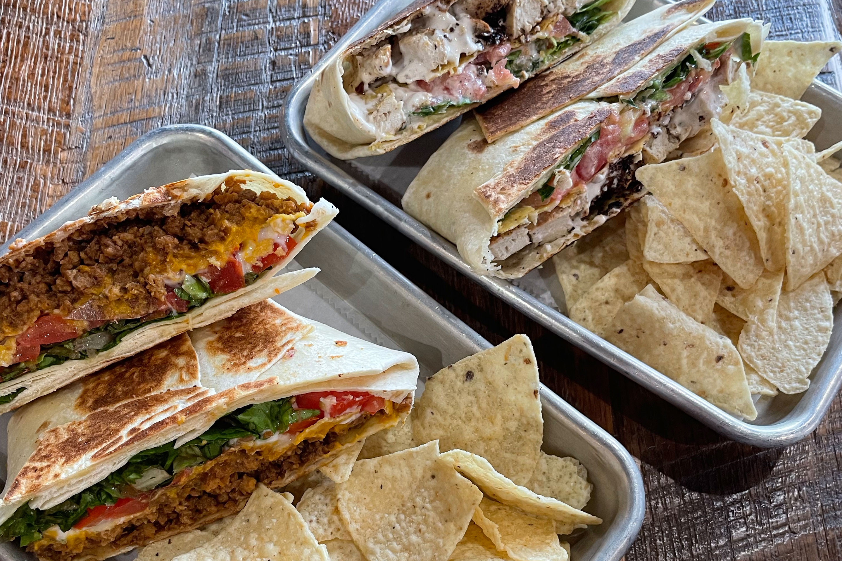 Two metal serving trays containing nacho chips and a specialty crunch, tortilla wrap on a wooden table.