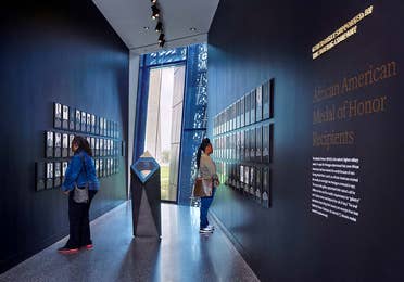Two women stand in a black hallway curated with photography, statues and supportive signage.