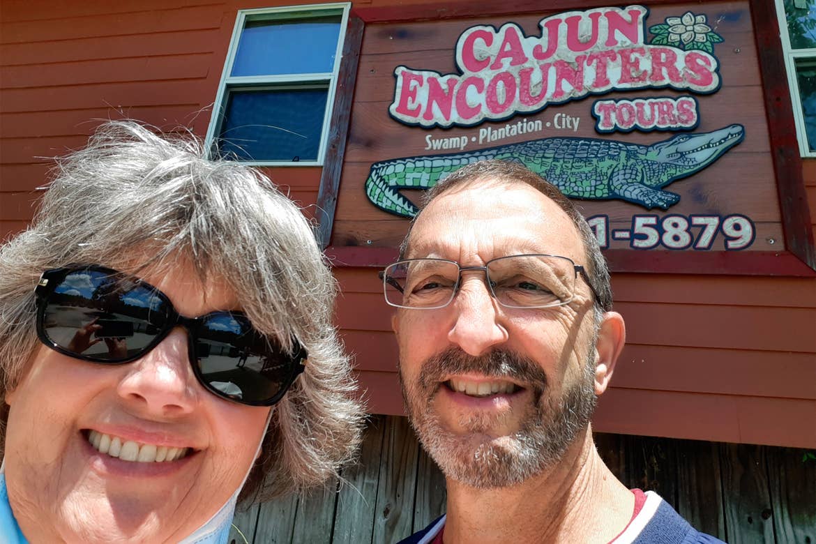 A woman wearing a safety mask and sunglasses stands next to a man in reading glasses under an exterior sign that reads 'Cajun Encounters Tours'.