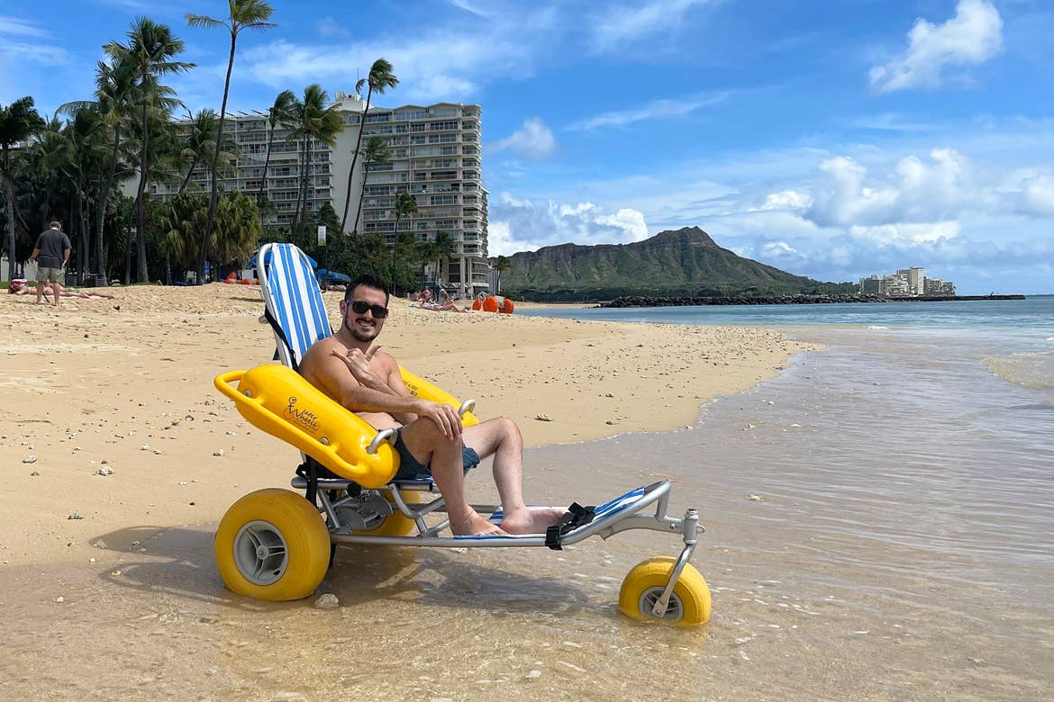 Featured Contributor, Danny Pitaluga, wears sunglasses and swim trunks while sitting in a yellow beach wheelchair on the sand as waves roll in.