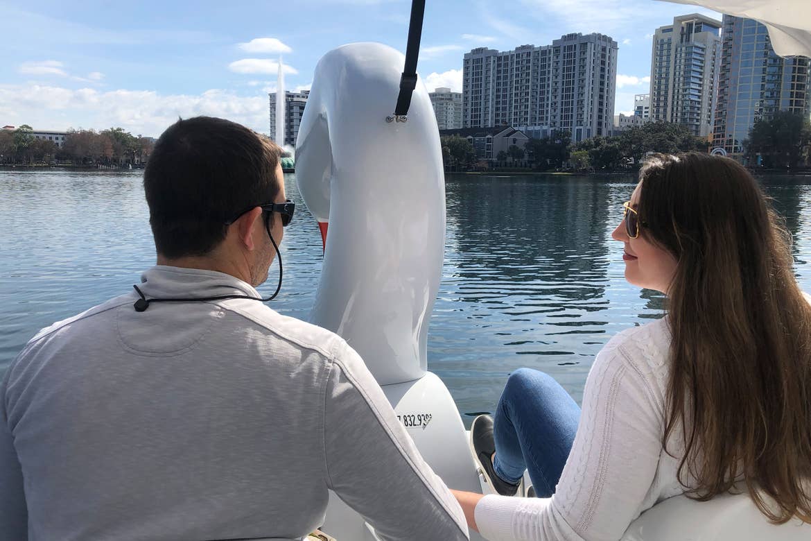 Featured contributor, Tori Ferrante (right) and her husband wear black sunglasses and white sweaters while paddling a white Swan boat in Lake Eola under a blue sky.