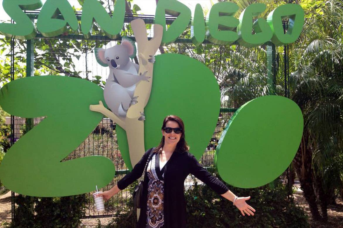 Jenn C. Harmon wears a black cardigan and patterned blouse in front of a green marquee that reads, 'San Diego Zoo' with a koala graphic.
