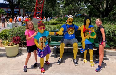 Two young girls (far left and far right) pose with a LEGO family on a bench.