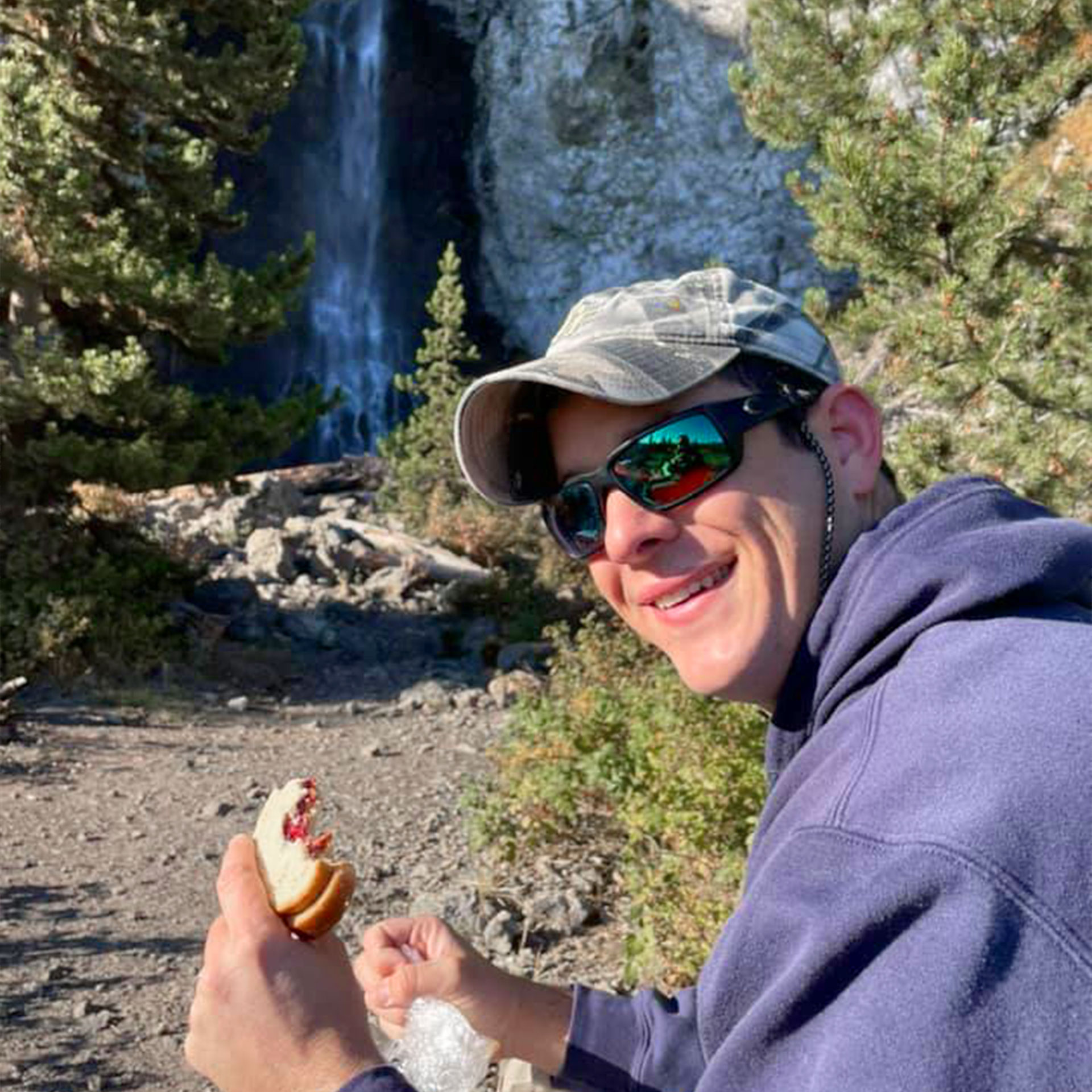 A man wearing a baseball cap, sunglasses and a blue hoodie holds a pb&j sandwich near a waterfall outdoors.
