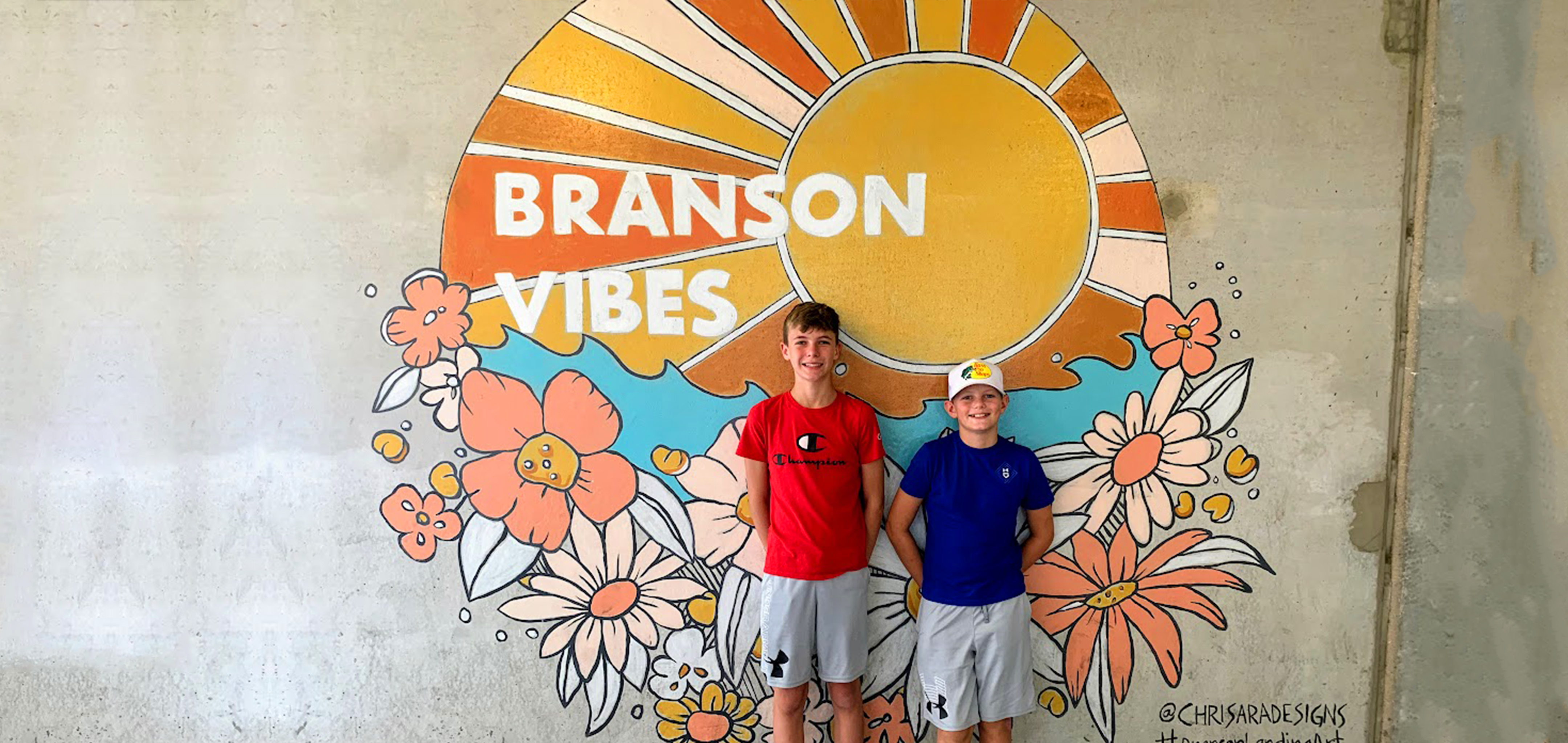 Two boys stand in front of a mural that reads,  'Branson Vibes'.