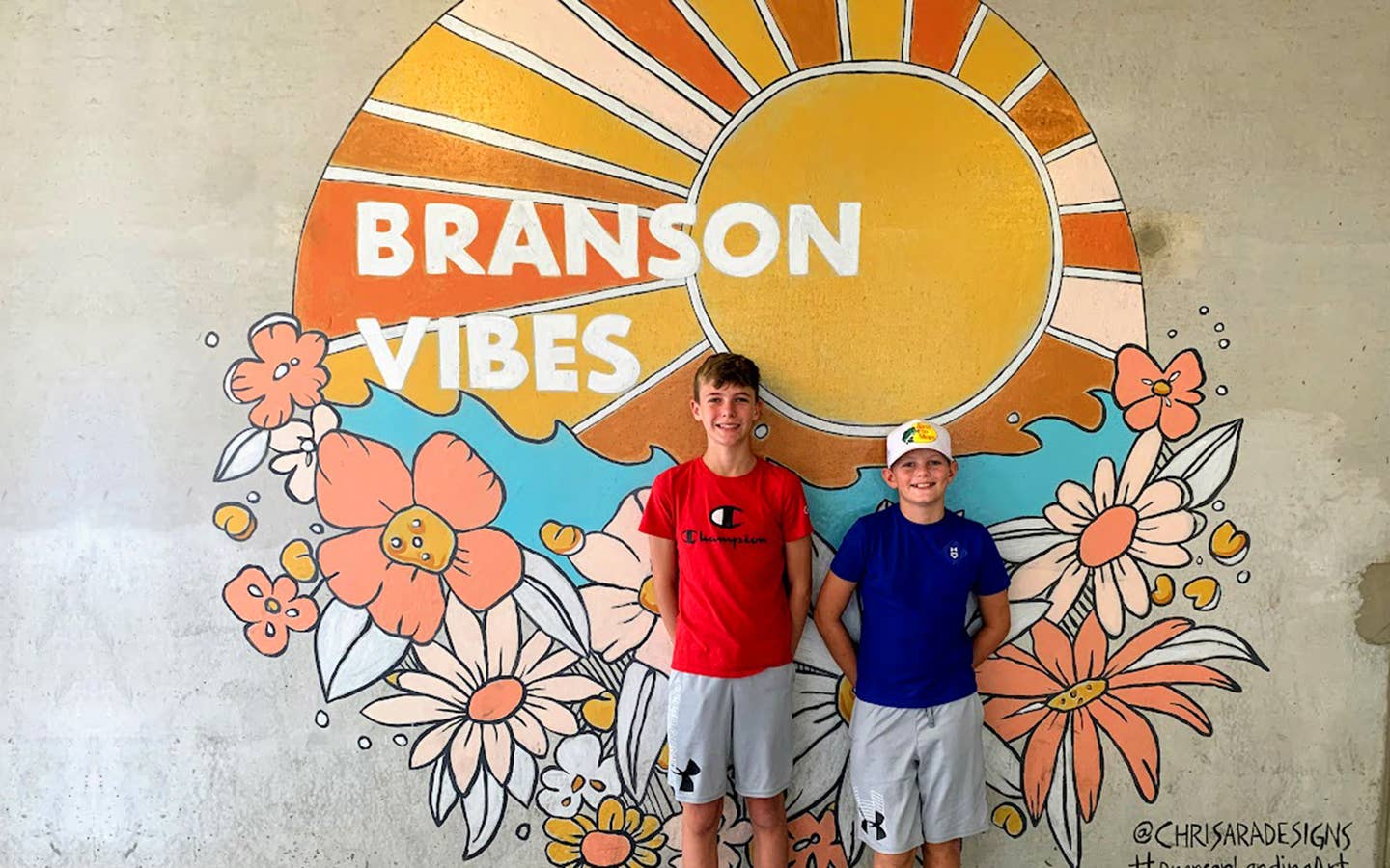 Two boys stand in front of a mural that reads,  'Branson Vibes'.
