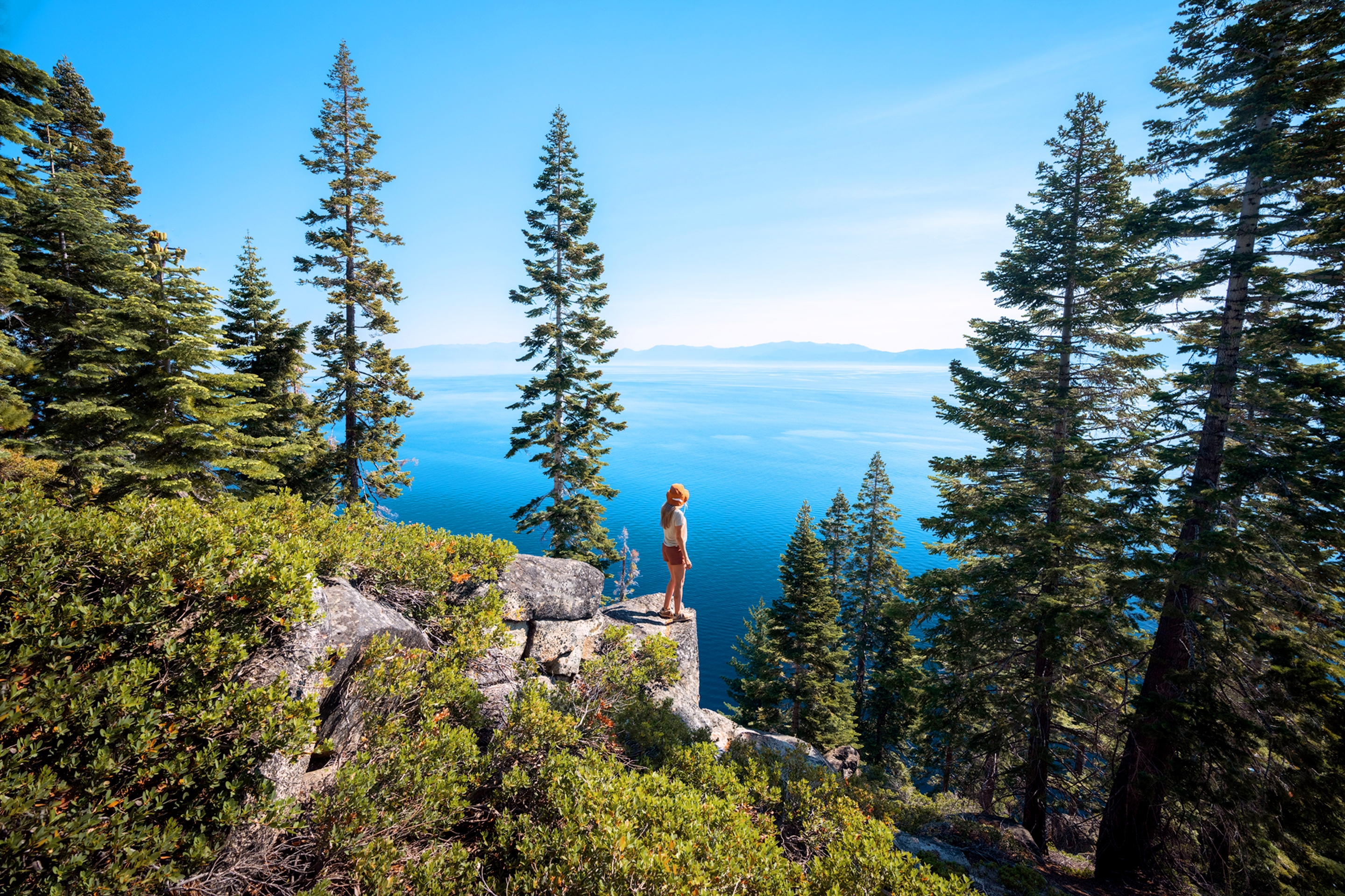 A caucasian woman wearing a white shirt, white baseball cap, shorts and boots sits on a rock at a high elevation overlooking a lake surrounded by pine trees on a mountain.