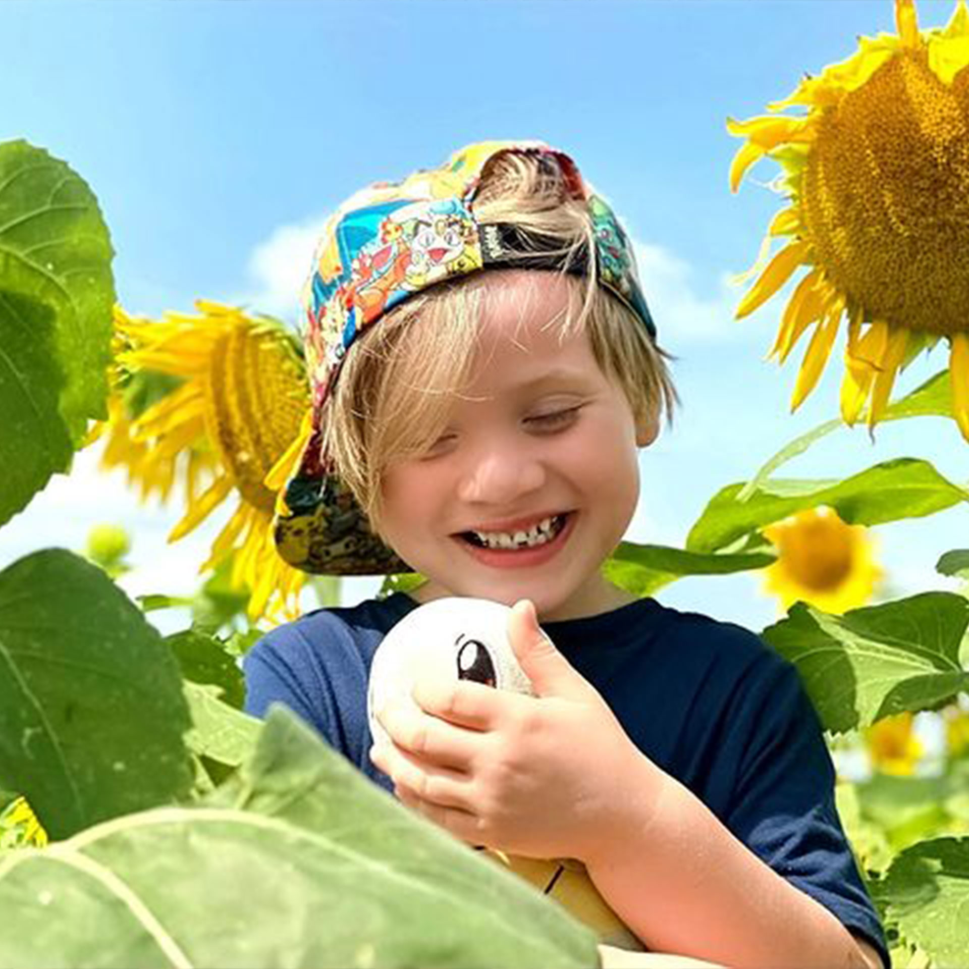 Featured Contributor, Alicia Trautwein's son stands in a sunflower field wearing a hat and holding a plush Squirtle.