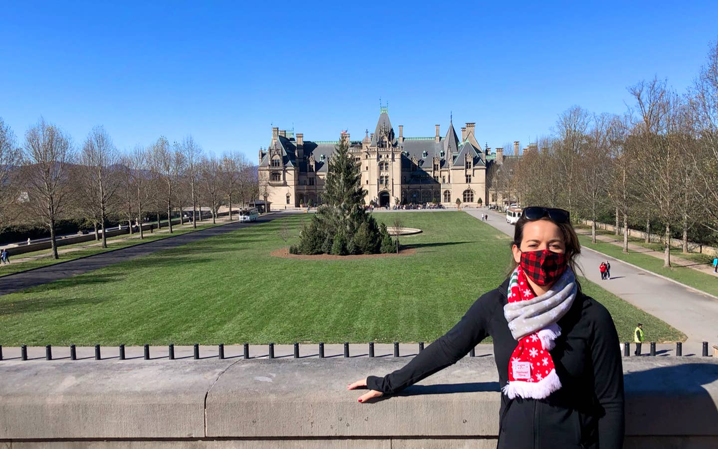 Author, Jenn C. Harmon, stands in front of the Biltmore Estate wearing a buffalo plaid red and black face mask and festive scarf over a black jacket.