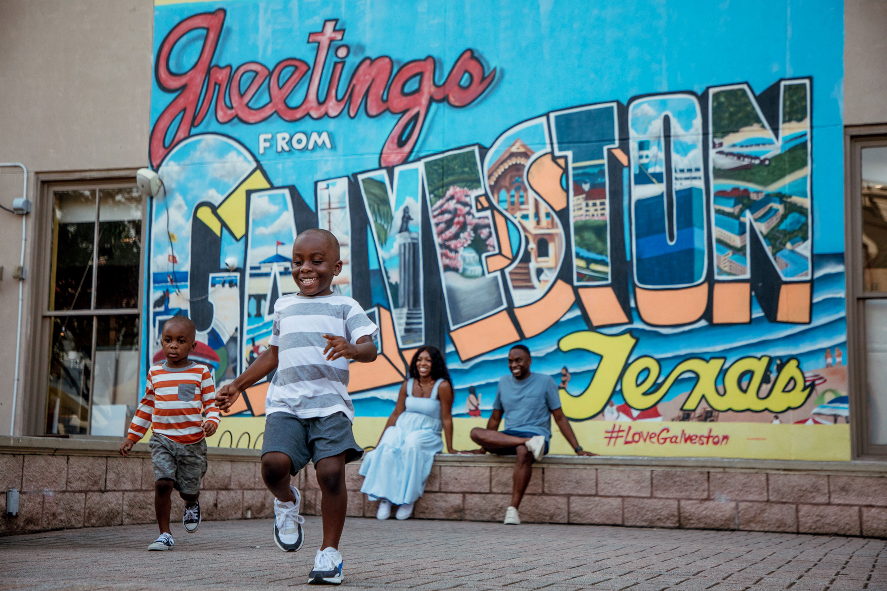 Two young boys running from a woman and man sitting under a walll mural that reads, 'Greetings from Galveston Texas.'