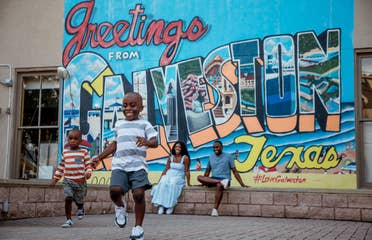 Two young boys running from a woman and man sitting under a walll mural that reads, 'Greetings from Galveston Texas.'