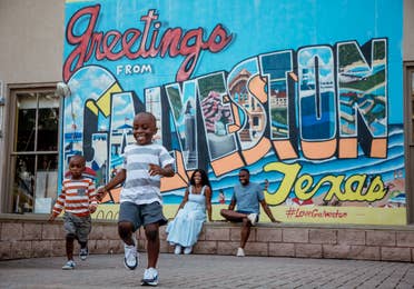 Two young boys running from a woman and man sitting under a walll mural that reads, 'Greetings from Galveston Texas.'