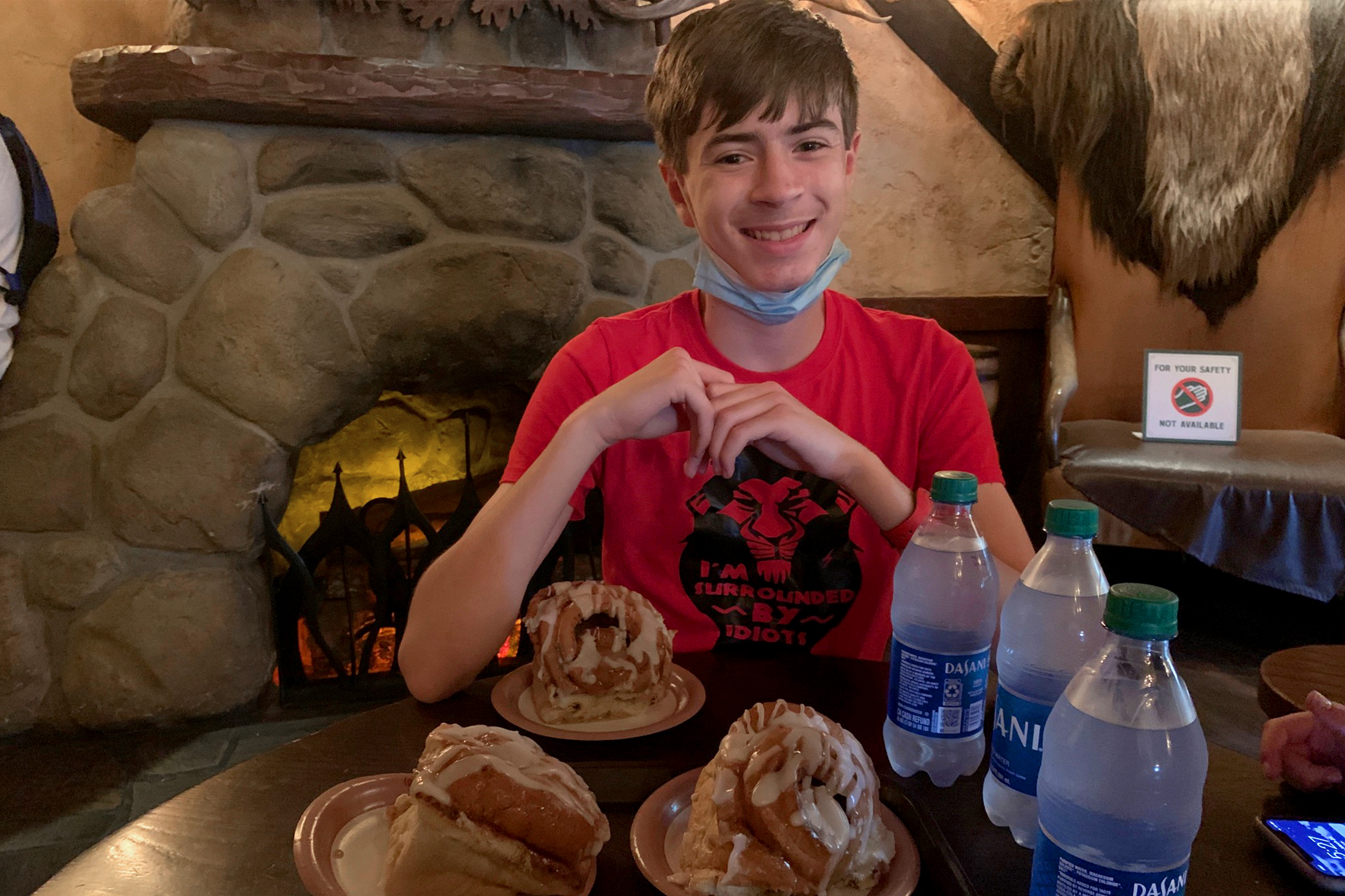A young man wearing a red t-shirt and a safety mask on his chin sits at a table surrounded by three Dasani water bottles and three cinnamon rolls inside a restaurant.