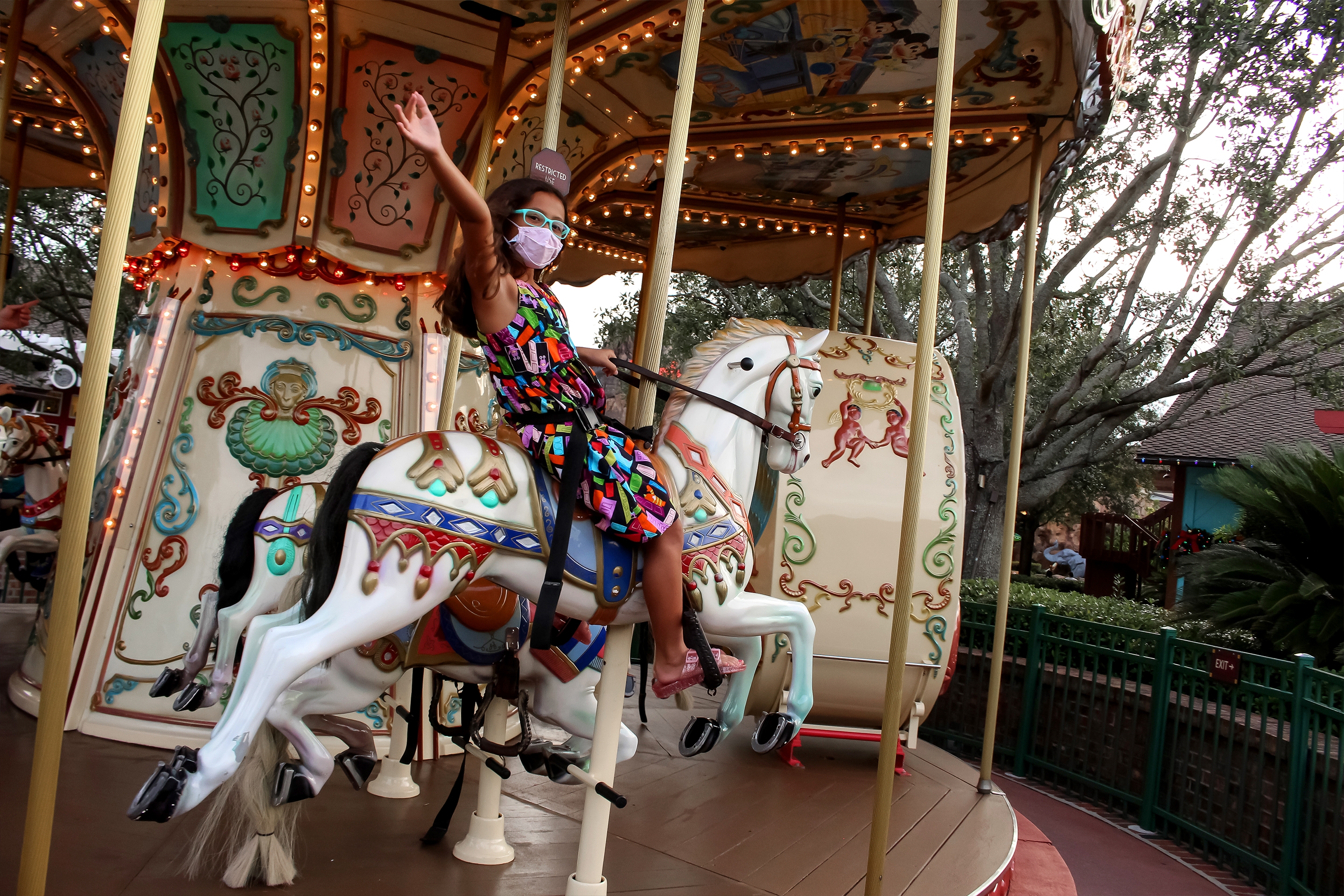 An Asian girl wears a multi-colored dress, blue glasses, and a safety mask while riding a carousel outdoors.