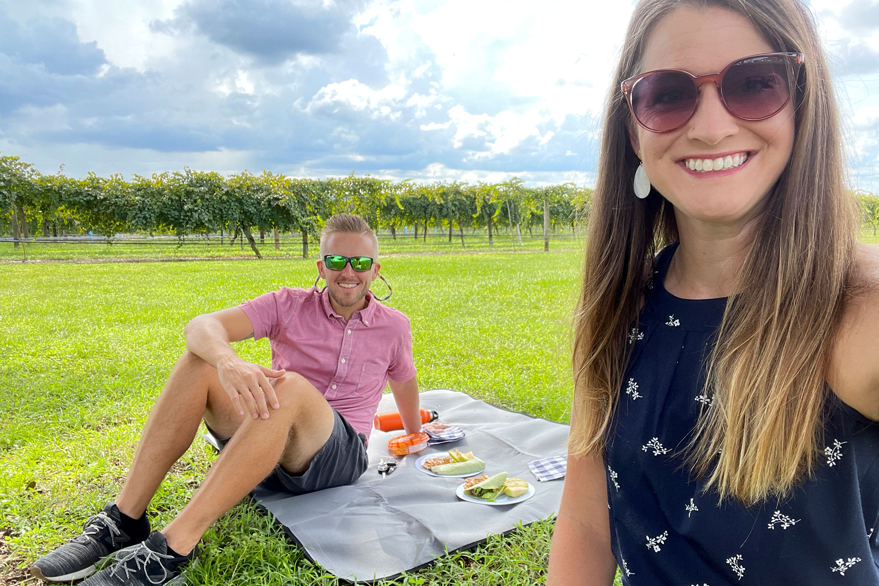 A man (left) wears a red button-up shirt and dark shorts while seated on a picnic blanket next to a woman (right) wearing a navy dress near a row of vineyard trees.