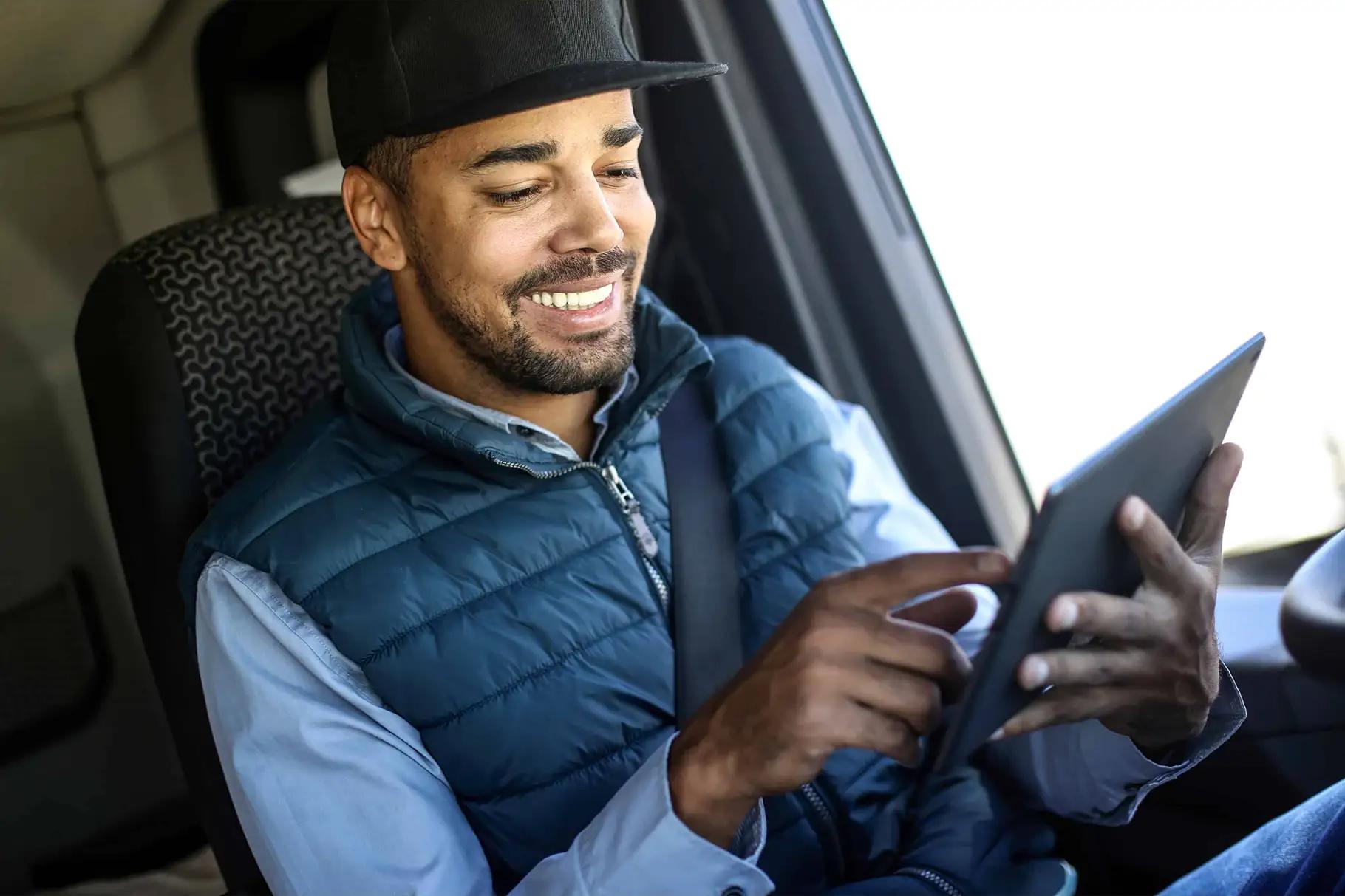 A smiling male truck driver wearing a black cap and blue quilted vest, seated in a vehicle cab with seatbelt fastened, using a tablet device
