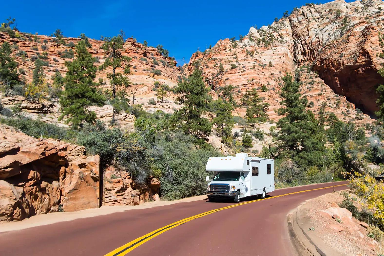 A white Class C motorhome RV driving along a red-paved two-lane road through a scenic red sandstone canyon with pine trees, characteristic of Zion National Park in Utah