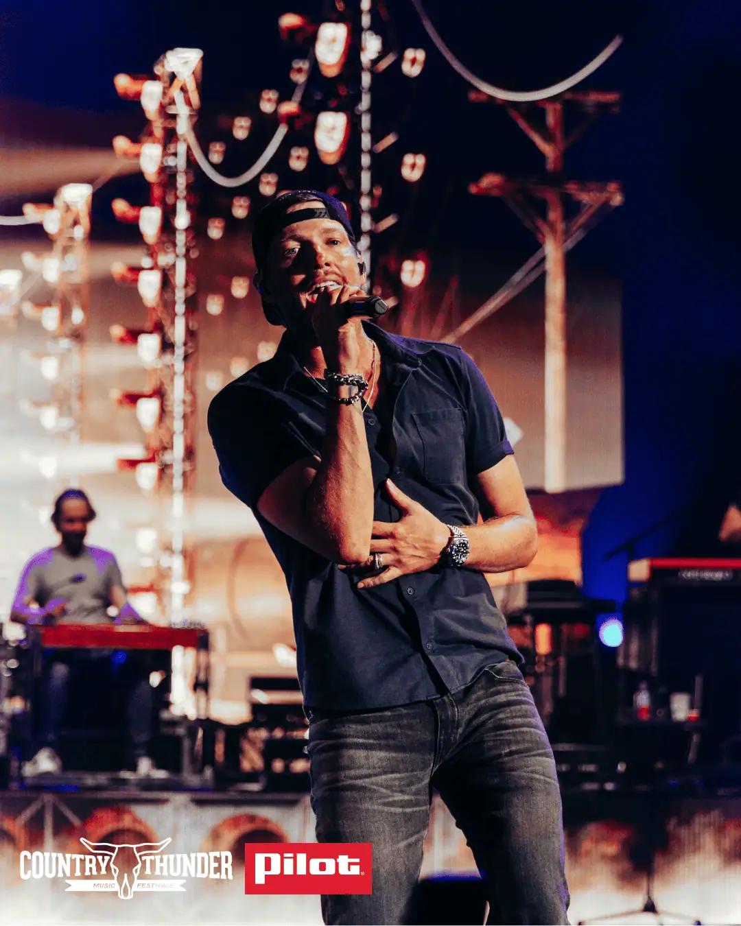 A male performer in a dark blue shirt and backwards cap sings into a handheld microphone on a concert stage at Country Thunder Music Festival, sponsored by Pilot, with warm stage lighting and a keyboard player visible in the background.
