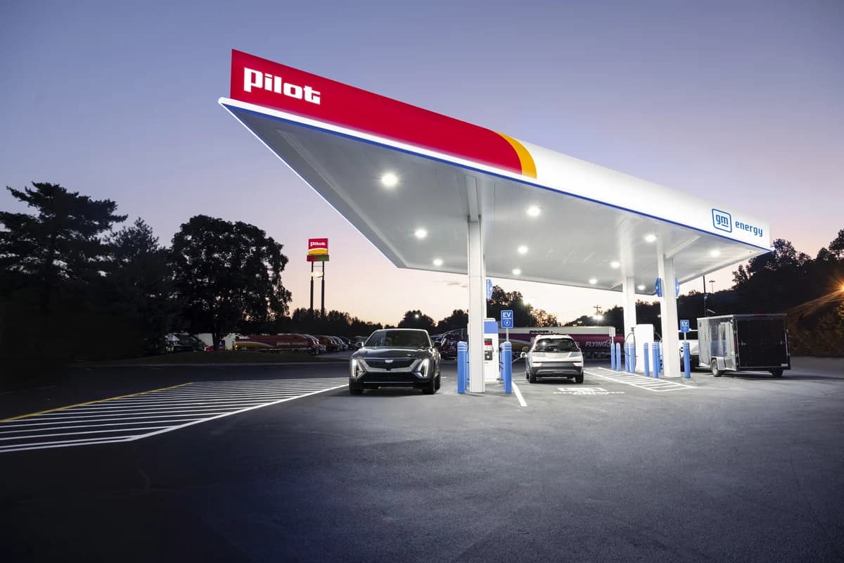 A Pilot EV charging station co-branded with GM Energy, featuring a large illuminated canopy with red, yellow, and blue branding at dusk, with two electric vehicles including a Cadillac plugged into blue EV charging stalls