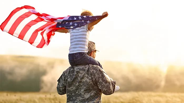 A child sits on the shoulders of a person wearing military camouflage, holding an American flag outstretched against a bright, sunlit field