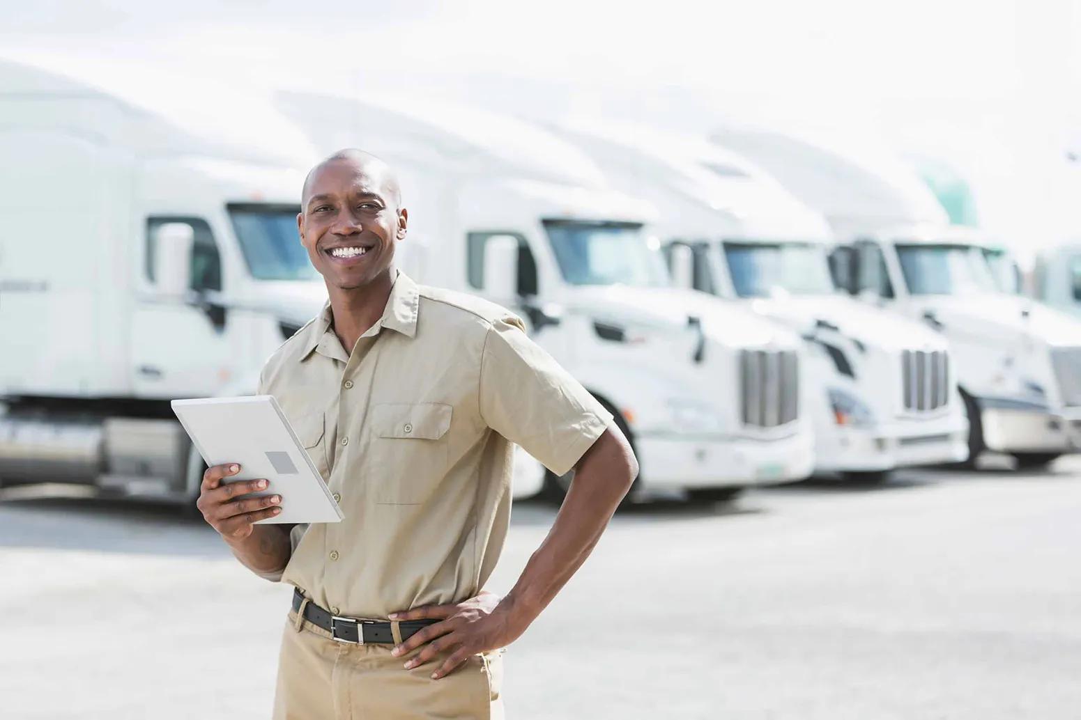 Smiling man in a tan short-sleeve shirt and khaki pants holding a tablet in one hand with the other hand on his hip, standing in front of a row of white semi-trucks in a parking lot.