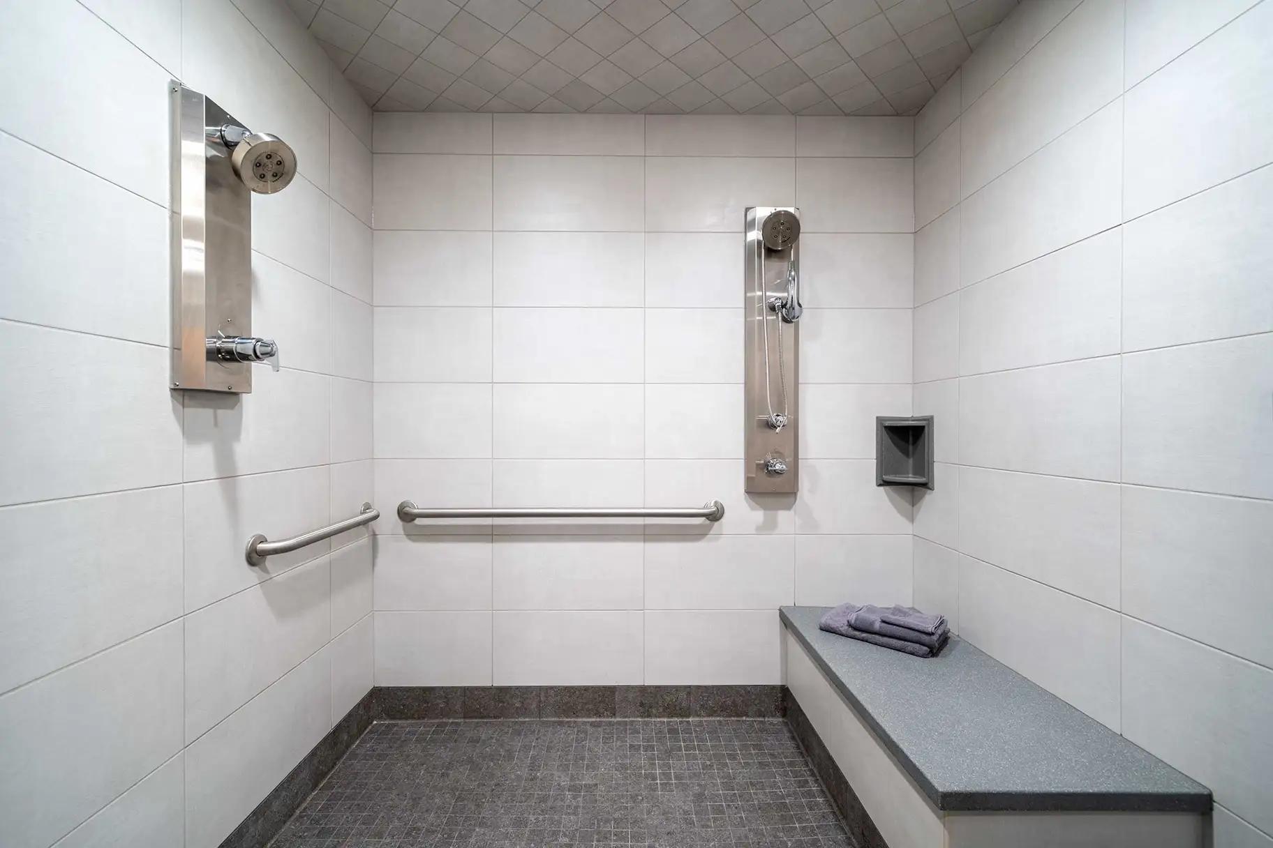 ADA-accessible roll-in shower with white tile walls, dark gray mosaic tile floor, built-in bench with folded towels, stainless steel grab bars, two shower fixtures including a handheld wand on a slide bar, and a recessed niche shelf